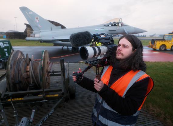 Behind-the-scenes image of a BFBS Creative videographer wearing a high-visibility vest and operating a professional cinema camera with a telephoto lens and microphone. A Royal Air Force Typhoon aircraft is parked on the airfield in the background during a recruitment filming session.