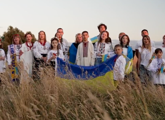 Group of Ukrainian adults and children wearing traditional embroidered clothing and holding Ukrainian flags in a field.