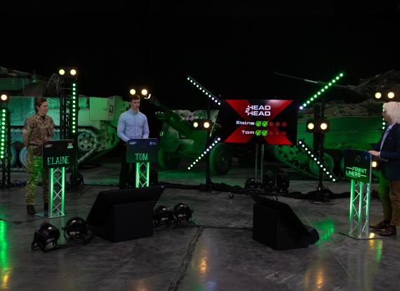 Two contestants and a presenter stand at illuminated podiums on a studio set, with military vehicles, green lighting and a game show scoreboard in the background.