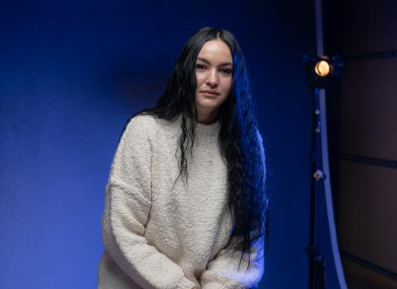 Portrait of a woman with long dark hair wearing a cream jumper, seated against a blue-lit studio backdrop with a studio light visible behind her.