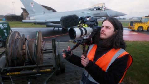 Behind-the-scenes image of a BFBS Creative videographer wearing a high-visibility vest and operating a professional cinema camera with a telephoto lens and microphone. A Royal Air Force Typhoon aircraft is parked on the airfield in the background during a recruitment filming session.