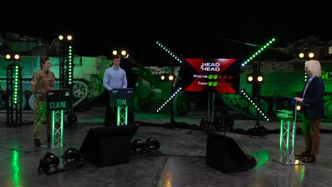 Two contestants and a presenter stand at illuminated podiums on a studio set, with military vehicles, green lighting and a game show scoreboard in the background.