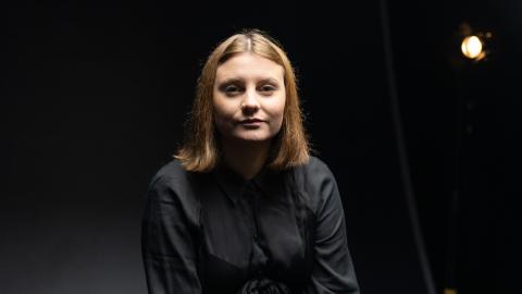 Woman wearing a black shirt seated for a portrait against a dark studio backdrop, with a studio light visible behind her.
