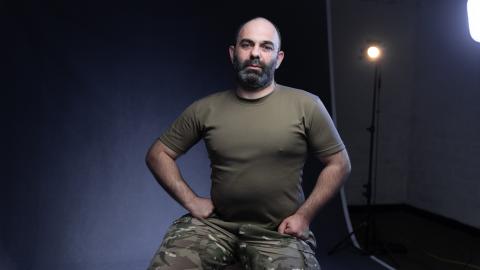 Man wearing an olive green T-shirt and camouflage trousers seated for a portrait against a dark studio backdrop, with production lighting visible behind him.