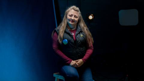 Woman wearing a black vest over a burgundy top seated for a portrait against a blue-lit studio backdrop, with a studio light visible behind her.