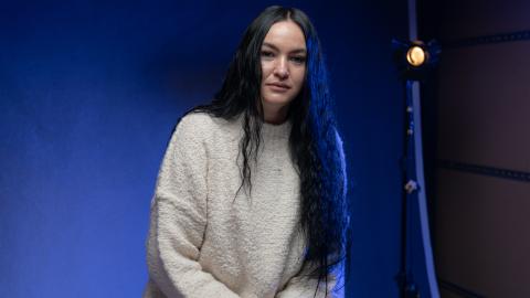 Portrait of a woman with long dark hair wearing a cream jumper, seated against a blue-lit studio backdrop with a studio light visible behind her.