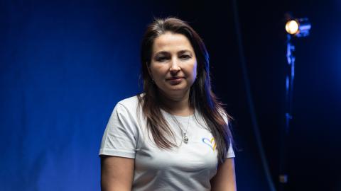 Woman wearing a white T-shirt seated for a portrait against a blue-lit studio backdrop, with a studio light visible behind her.