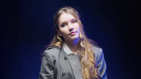 Portrait of a woman with long blonde hair wearing a grey jacket, seated against a dark blue-lit studio backdrop.