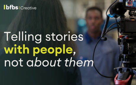 A banner graphic featuring a blurred behind-the-scenes interview and filming setup, with a camera in focus on the right-hand side. Overlaid text reads: “telling stories with people, not about them”. The design uses dark green gradients and subtle textured overlays in an editorial style associated with The Comms Dispatch by BFBS Creative.