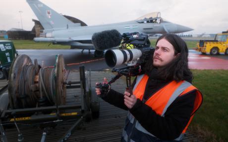 Behind-the-scenes image of a BFBS Creative videographer wearing a high-visibility vest and operating a professional cinema camera with a telephoto lens and microphone. A Royal Air Force Typhoon aircraft is parked on the airfield in the background during a recruitment filming session.