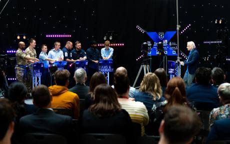 Live game show set with teams of military personnel at illuminated podiums, a presenter and seated audience, surrounded by cameras and colourful studio lighting.