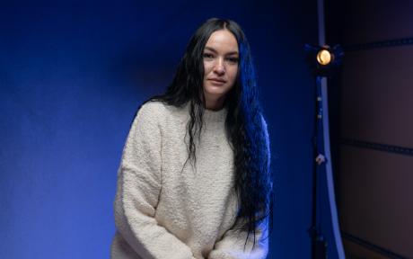Portrait of a woman with long dark hair wearing a cream jumper, seated against a blue-lit studio backdrop with a studio light visible behind her.