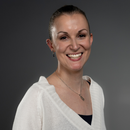 A woman smiles warmly at the camera. She has her hair pulled back and is wearing a white jumper, earrings and a necklace. The background is grey and softly lit.