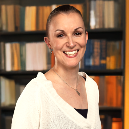 A woman smiles warmly at the camera. She has her hair pulled back and is wearing a white jumper, earrings and a necklace. The background is a blurred bookcase full of books. 
